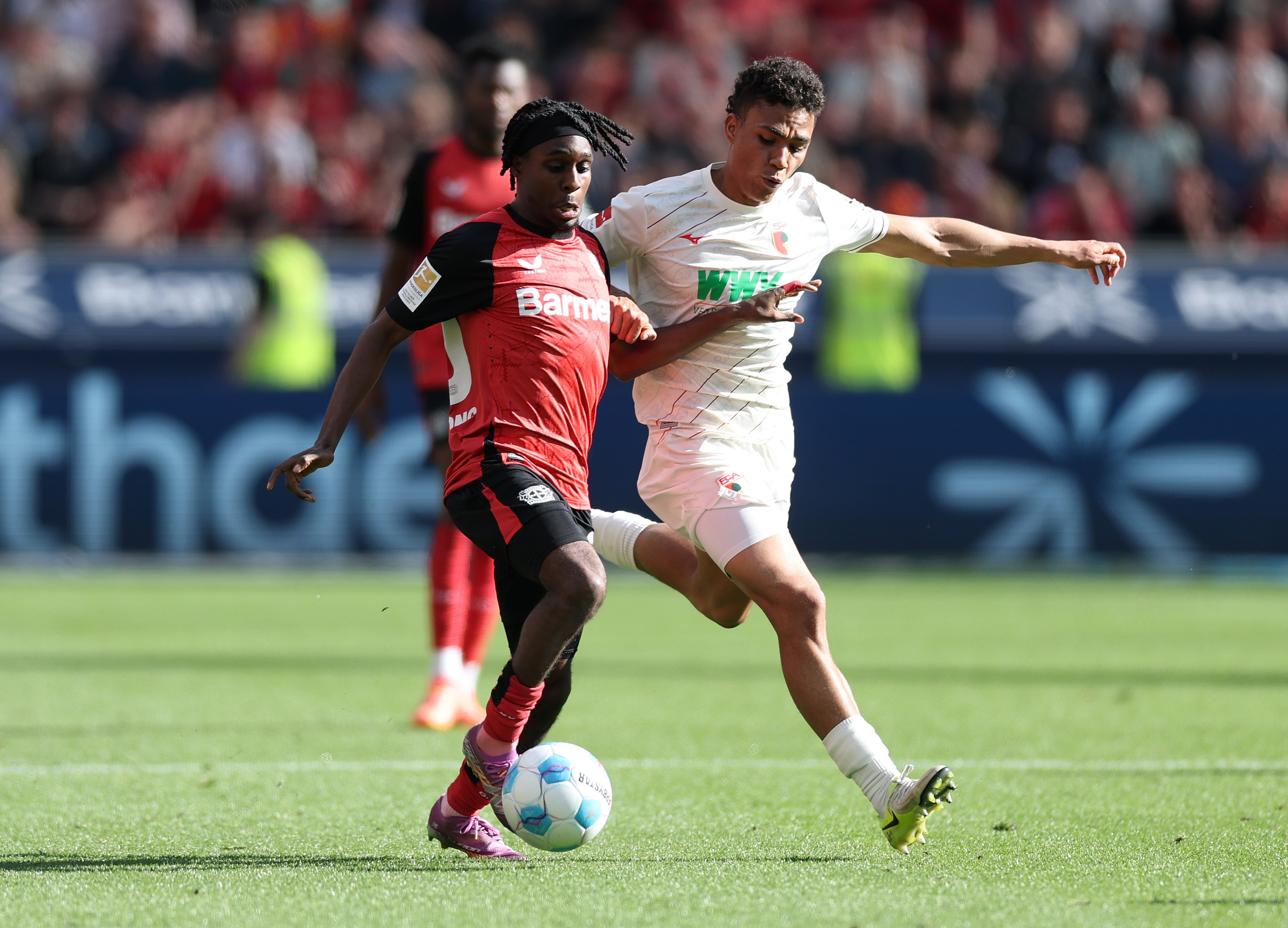 Jeremie Frimpong holds off an FC Augsburg player with his arm whilst dribbling with the ball.
